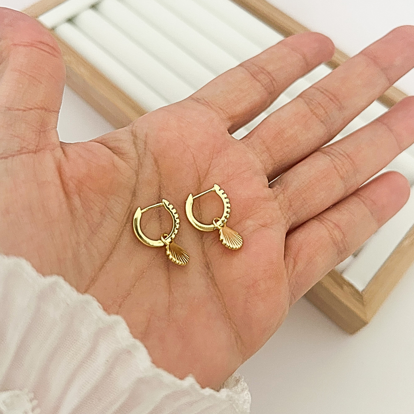 Pair of gold huggie hoop earrings with seashell charm held in open palm