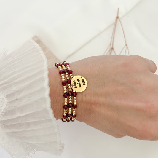 Elegant three-strand bracelet featuring deep burgundy crystal beads, gold-tone accents, and a round charm engraved with “Amar, Soñar y Reír,” worn on the wrist with a delicate lace sleeve.