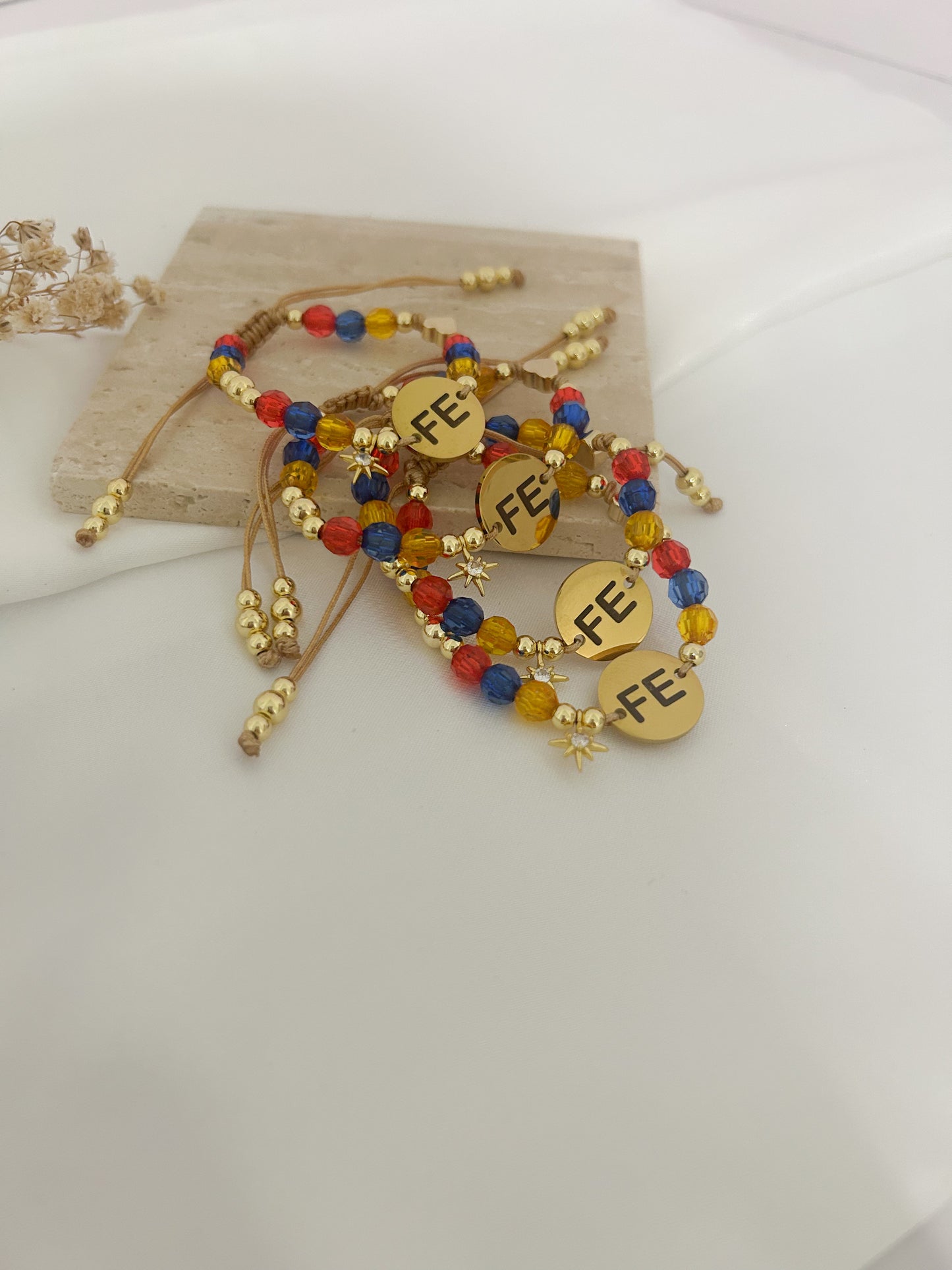 A set of gold-toned beaded bracelets with colorful red, blue, and yellow beads, featuring round gold charms engraved with 'FE' and small gold star charms, displayed on a light wood block over a white background.