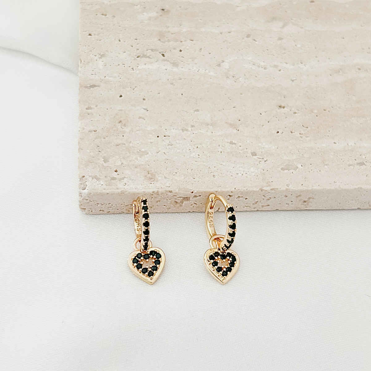 A pair of gold hoop earrings with dangling heart charms, decorated with small black stones, displayed on a white surface next to a beige stone block.