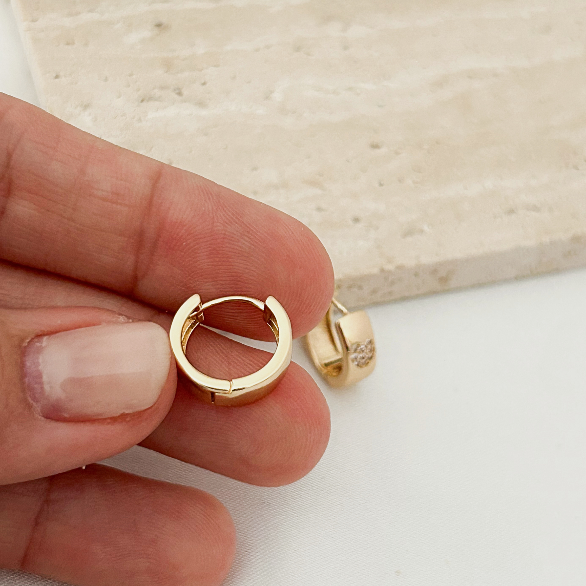 A close-up of a hand holding a small gold huggie hoop earring, with another matching earring resting on a light surface in the background.