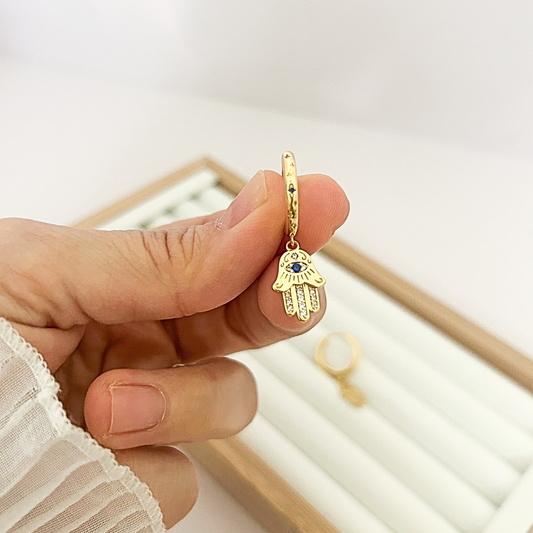 18K gold huggie hoop with Hamsa hand blue evil eye charm held in hand — close-up