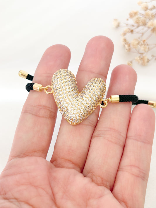 Gold heart-shaped bracelet with black cord held between fingers on a white background