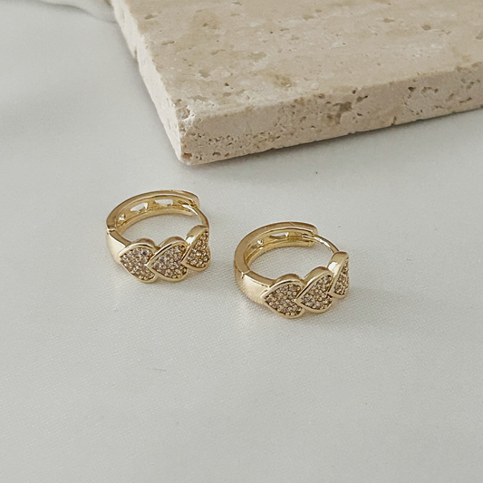 A pair of gold huggie hoop earrings with pavé-set clear stones in a wing-like design, displayed on a light gray surface with a beige stone prop in the background.