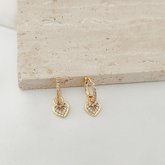 A pair of gold hoop earrings with dangling heart charms paved with small clear stones, displayed on a light beige stone slab over a white surface.