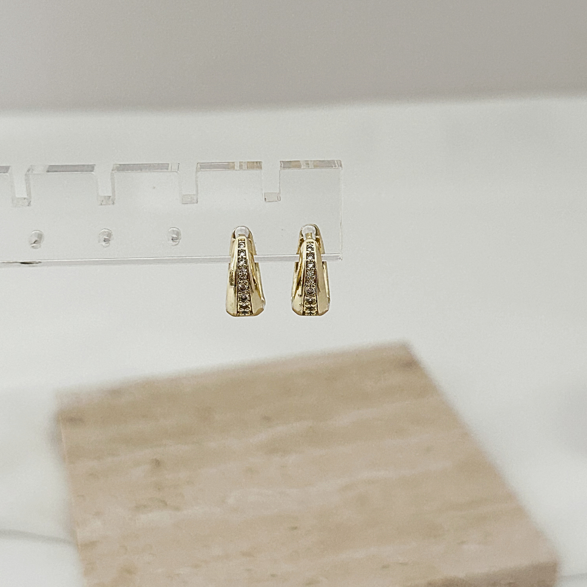 A pair of small gold hoop earrings with clear stones, displayed on a clear stand against a soft white background with a beige stone tile in the foreground.