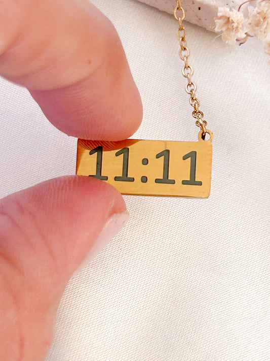 Gold necklace with '11:11' pendant held by a hand against a light background