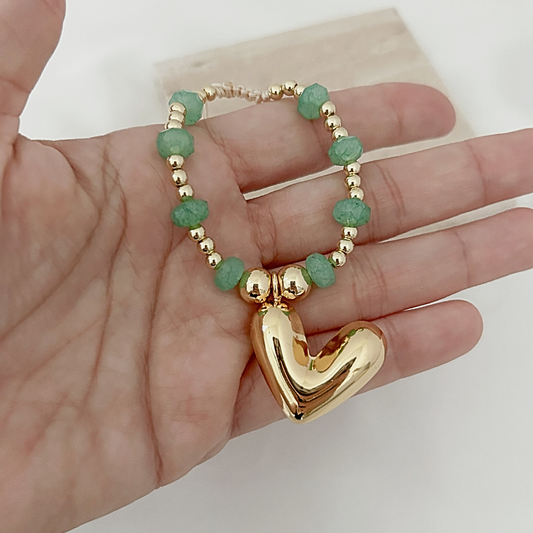 A gold heart-shaped pendant charm bracelet with green faceted beads and gold spacer beads, held in a person's hand.