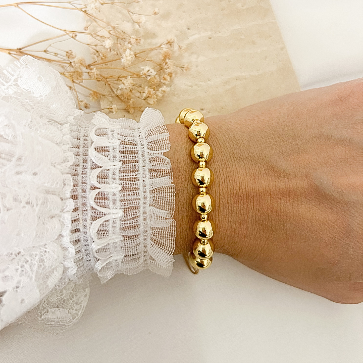 A gold beaded bracelet worn on a person's wrist, shown next to a white lace sleeve, with dried flowers in the background.
