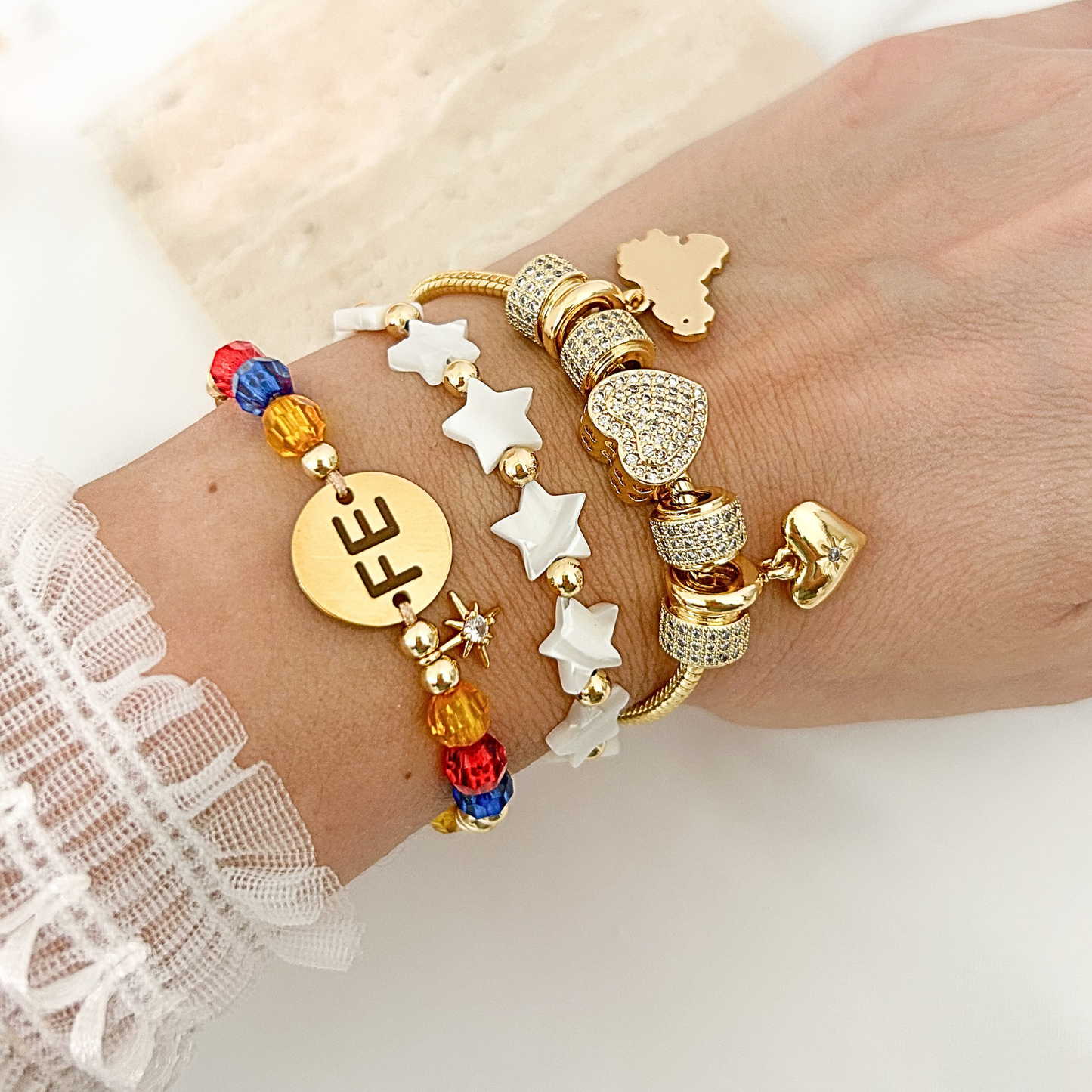 Layered bracelet stack featuring a Venezuela bracelet with colorful crystal beads, white star beaded bracelet, and gold charm bracelet with pavé heart accents. Elegant everyday jewelry designed for stacking.