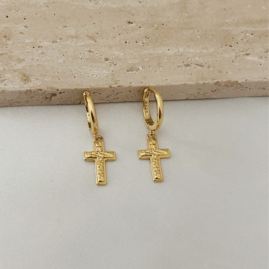 A pair of gold hoop earrings with small cross charms, displayed on a light gray surface with a beige stone slab in the background.