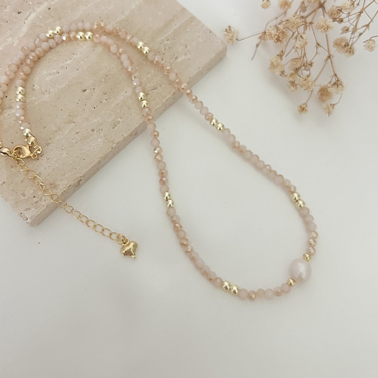 Handmade necklace with soft pink crystal beads, gold-tone spacers, and a natural freshwater pearl, displayed on a neutral stone background.