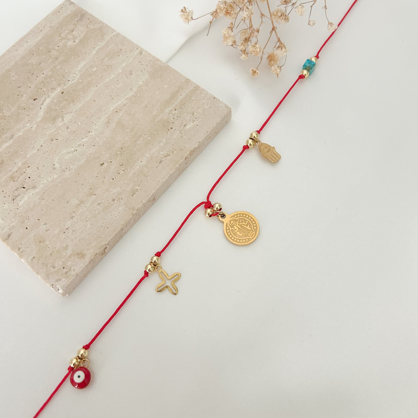 Close-up of a red cord necklace with meaningful protection symbols, including a Saint Benedict medal, Hamsa hand, evil eye charm, and gold-tone details.