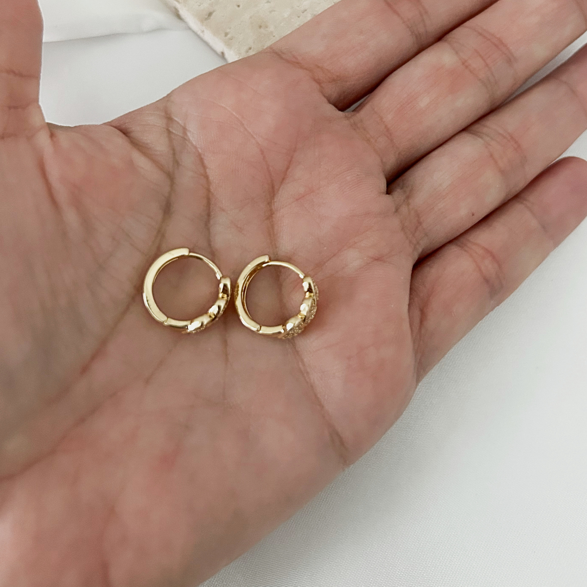 A pair of small gold hoop earrings resting on an open hand, shown against a light neutral background.