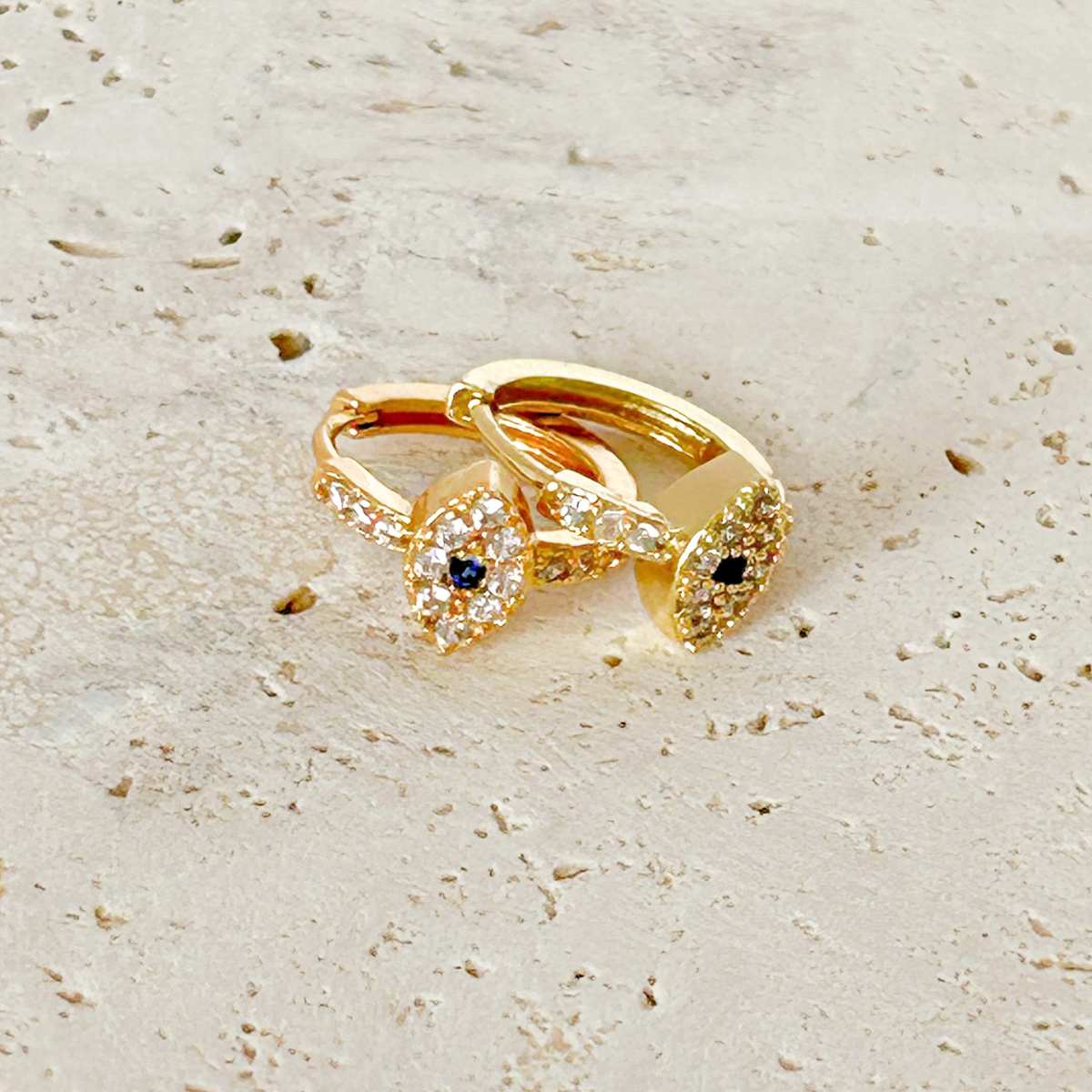 Close-up of gold evil eye hoop earrings with crystal details on a neutral stone background.