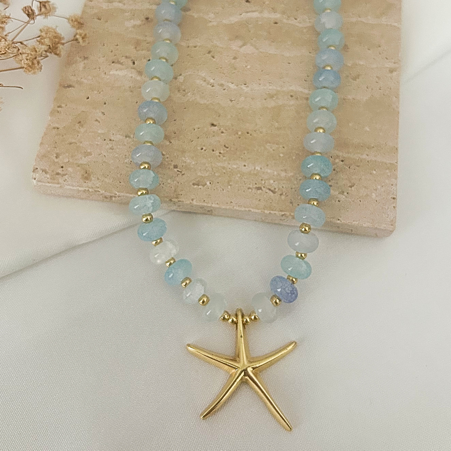 A beaded necklace with light blue and white round beads separated by small gold spacer beads, featuring a polished gold starfish pendant, displayed on a beige stone slab over a white fabric background.