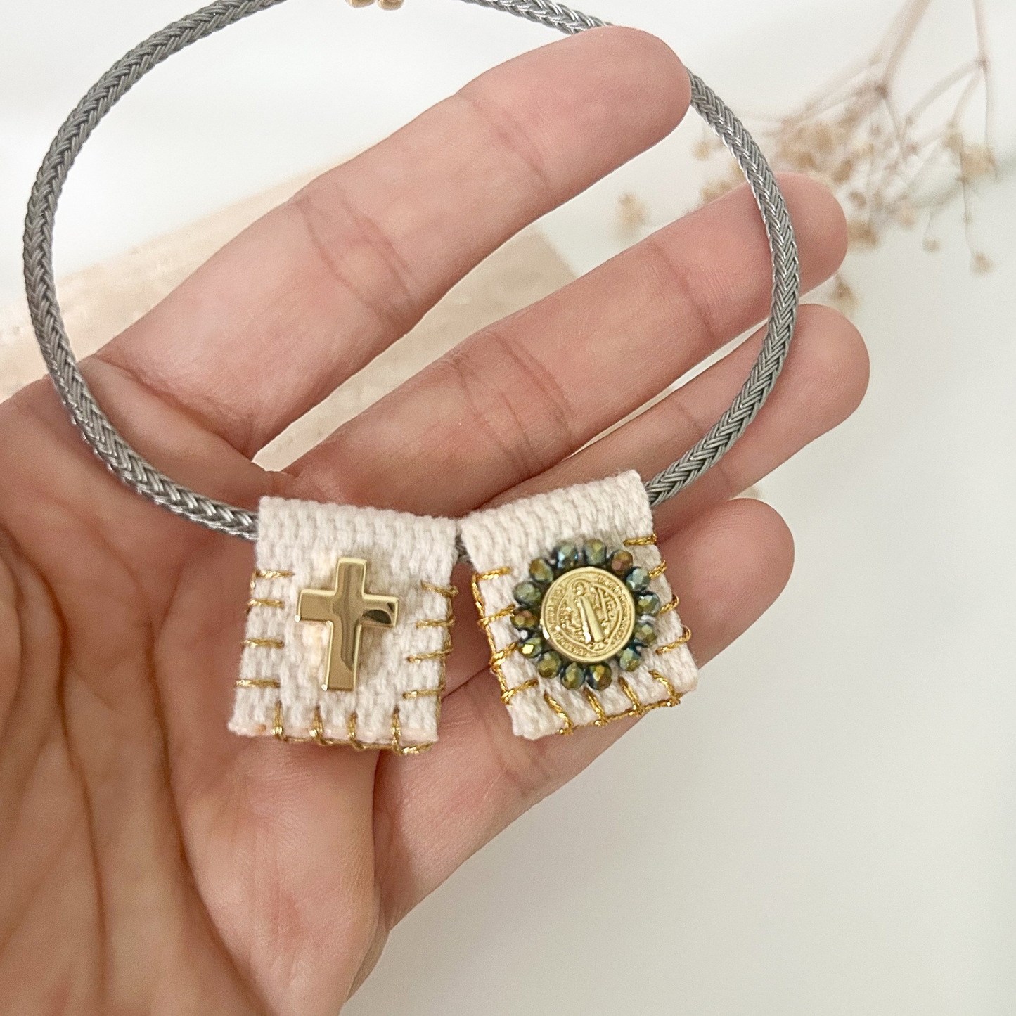 Close-up of a religious bracelet with textured ivory fabric details, a polished gold cross, Saint Benedict medal, and sparkling crystal accents on a gray cord band.