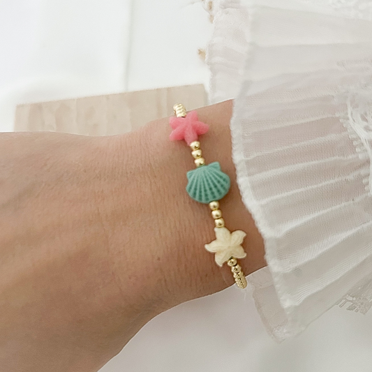 Delicate coastal-inspired bracelet worn on the wrist, designed with gold beads and colorful ocean charms including a pink starfish, aqua seashell, and ivory starfish, paired with a white ruffled sleeve.