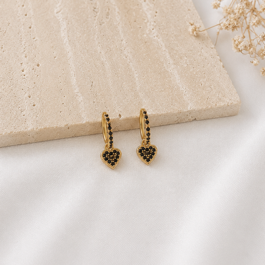 Pair of gold-tone huggie hoop earrings with black pavé crystal detailing and dangling black heart charms. Modern heart earrings with a bold yet elegant design, perfect for everyday wear, date nights, or layering with other gold jewelry.
