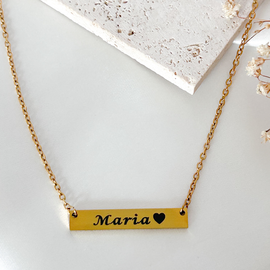 A gold bar necklace with the name 'Maria' and a small black heart engraved on the center, displayed on a light surface with a textured stone prop.