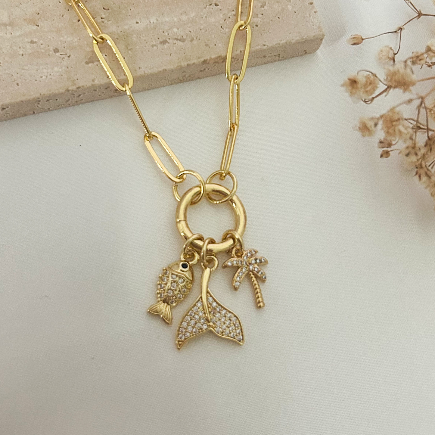 Elegant gold-tone paperclip necklace with tropical charms including a fish, palm tree, and mermaid tail, styled on a soft neutral backdrop for a coastal-inspired look.