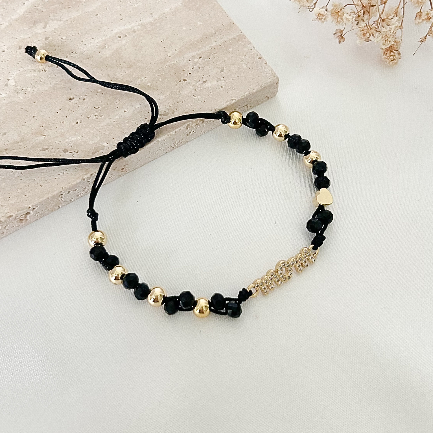 Adjustable black and gold bracelet displayed on a stone tray with dried florals, featuring faceted black beads, polished gold spacers, a tiny heart accent, and a glittering “Mom” centerpiece.