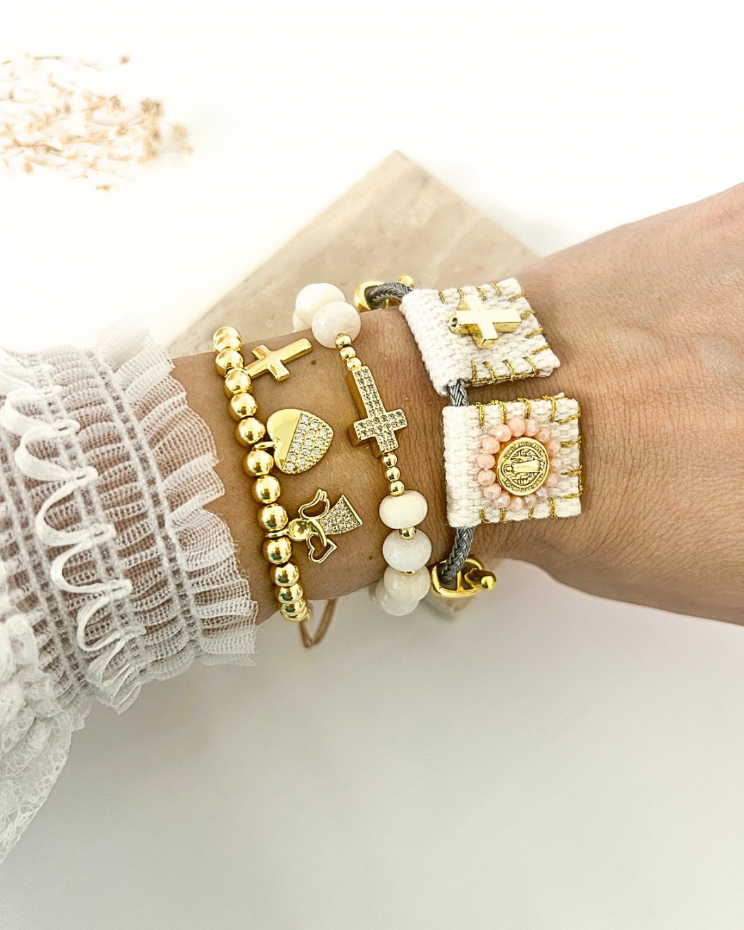 Layered gold, white, and blush bracelet with cross, heart, angel, and religious medal charms style on a wrist.