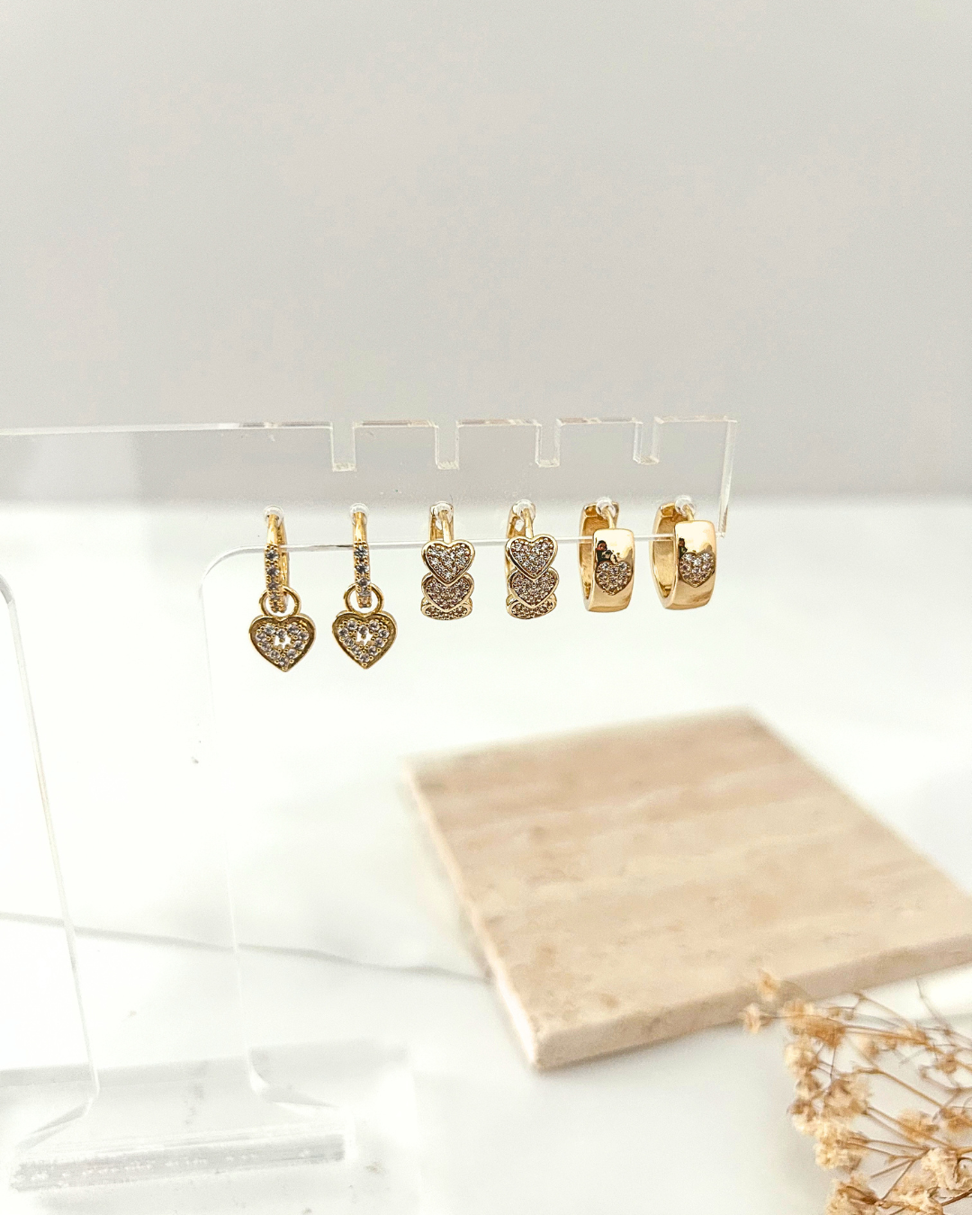 Three pairs of gold heart earrings with crystal details displayed on a clear acrylic jewelry stand.