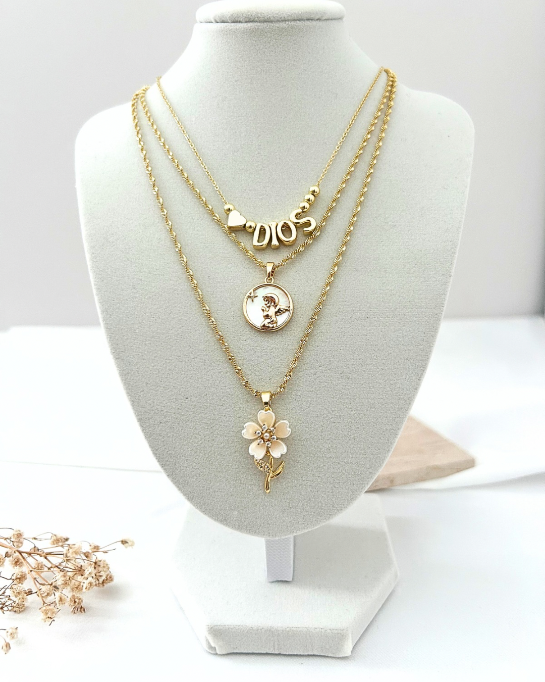 Layered gold necklaces with faith-inspired charms, including a "Dios" necklace, round angel pendant, and floral pendant displayed on a white jewelry bust.