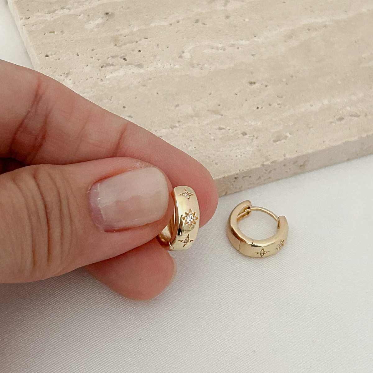 A close-up of a hand holding a small gold huggie hoop earring, with another matching gold hoop earring placed beside it on a white surface.