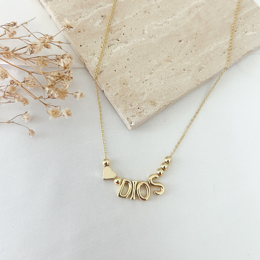 A gold necklace with a 'DIOS' nameplate pendant and a small heart charm, displayed on a light surface next to a beige stone tile and dried flowers.