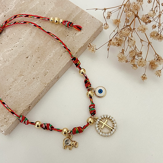 Handmade protection bracelet with colorful braided cord, gold beads, crystal cross pendant, evil eye detail, and elephant charm, displayed on a neutral stone surface with dried flowers.