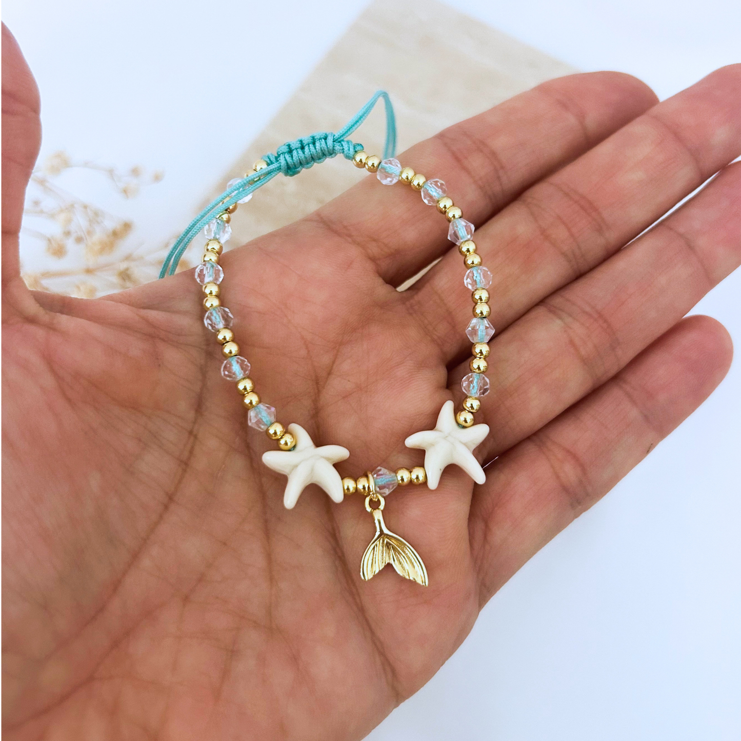 Close-up of an adjustable turquoise cord bracelet with white starfish beads, faceted crystal beads, gold-tone accents, and a dangling mermaid tail charm. Lightweight and ideal for everyday beach style.