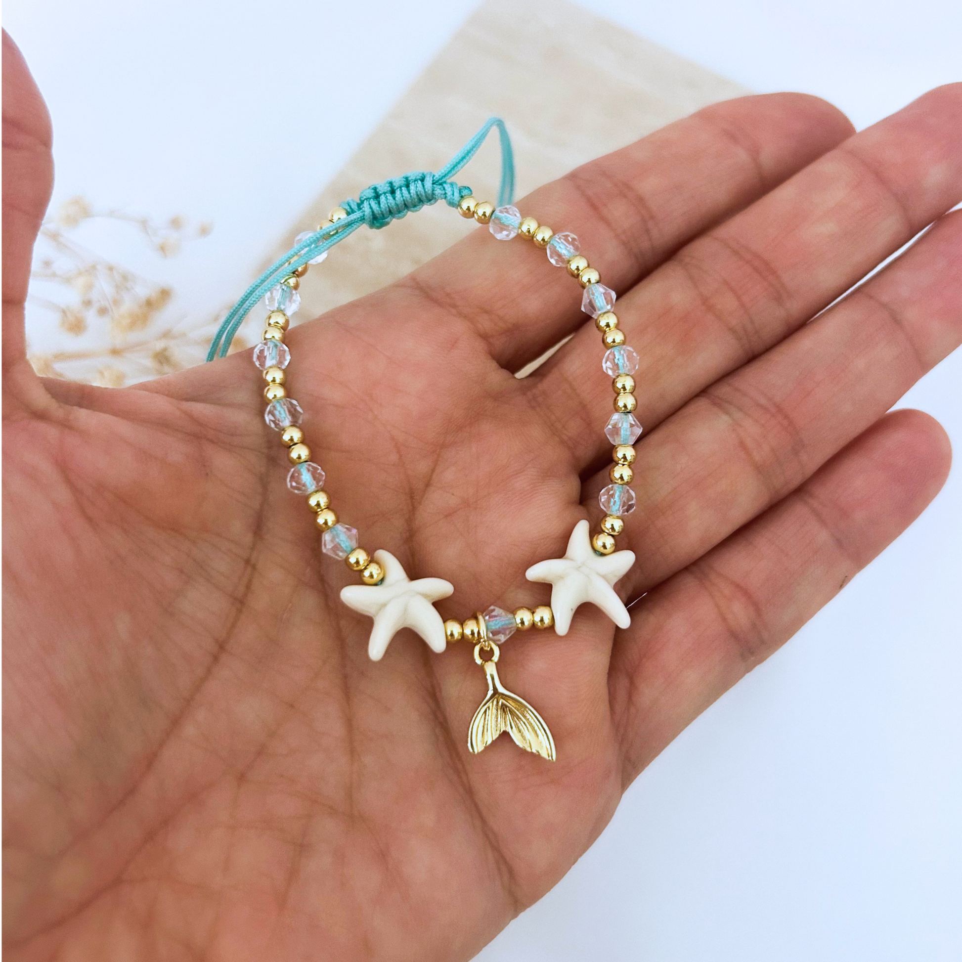 Close-up of an adjustable turquoise cord bracelet with white starfish beads, faceted crystal beads, gold-tone accents, and a dangling mermaid tail charm. Lightweight and ideal for everyday beach style.