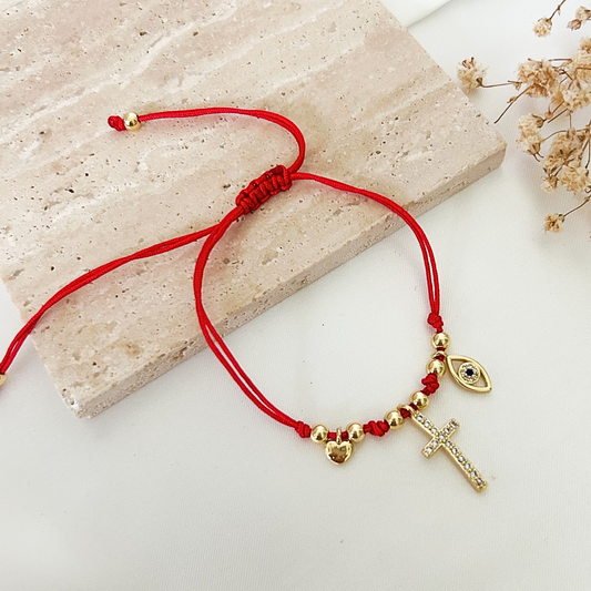 Adjustable red string bracelet with gold-tone beads and symbolic charms, featuring a crystal cross, evil eye charm, and small heart accent, displayed on a neutral stone surface with dried flowers.