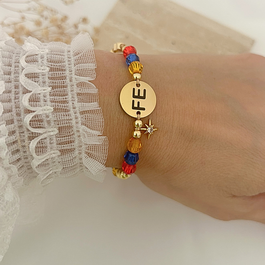 A close-up of a person's wrist wearing a colorful beaded bracelet with a round gold disc engraved with 'FE' and small gold star charms. The bracelet features red, blue, and gold beads, styled with a white lace sleeve.