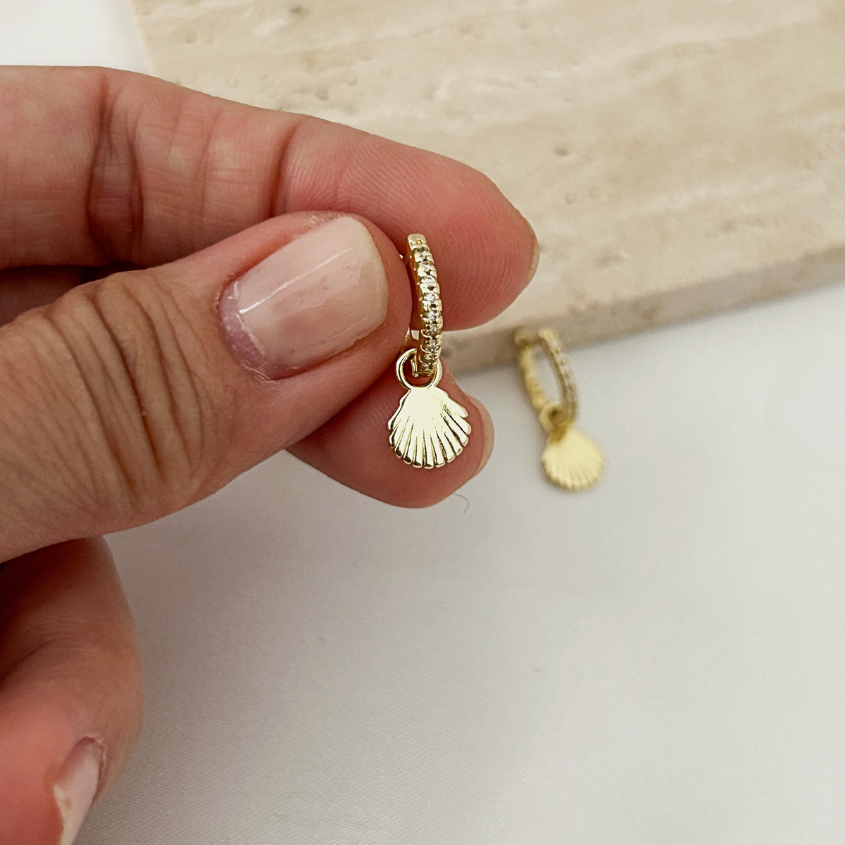 A close-up of a hand holding a small gold huggie hoop earring with a dangling scallop shell charm, with a second matching earring resting on a light surface in the background.