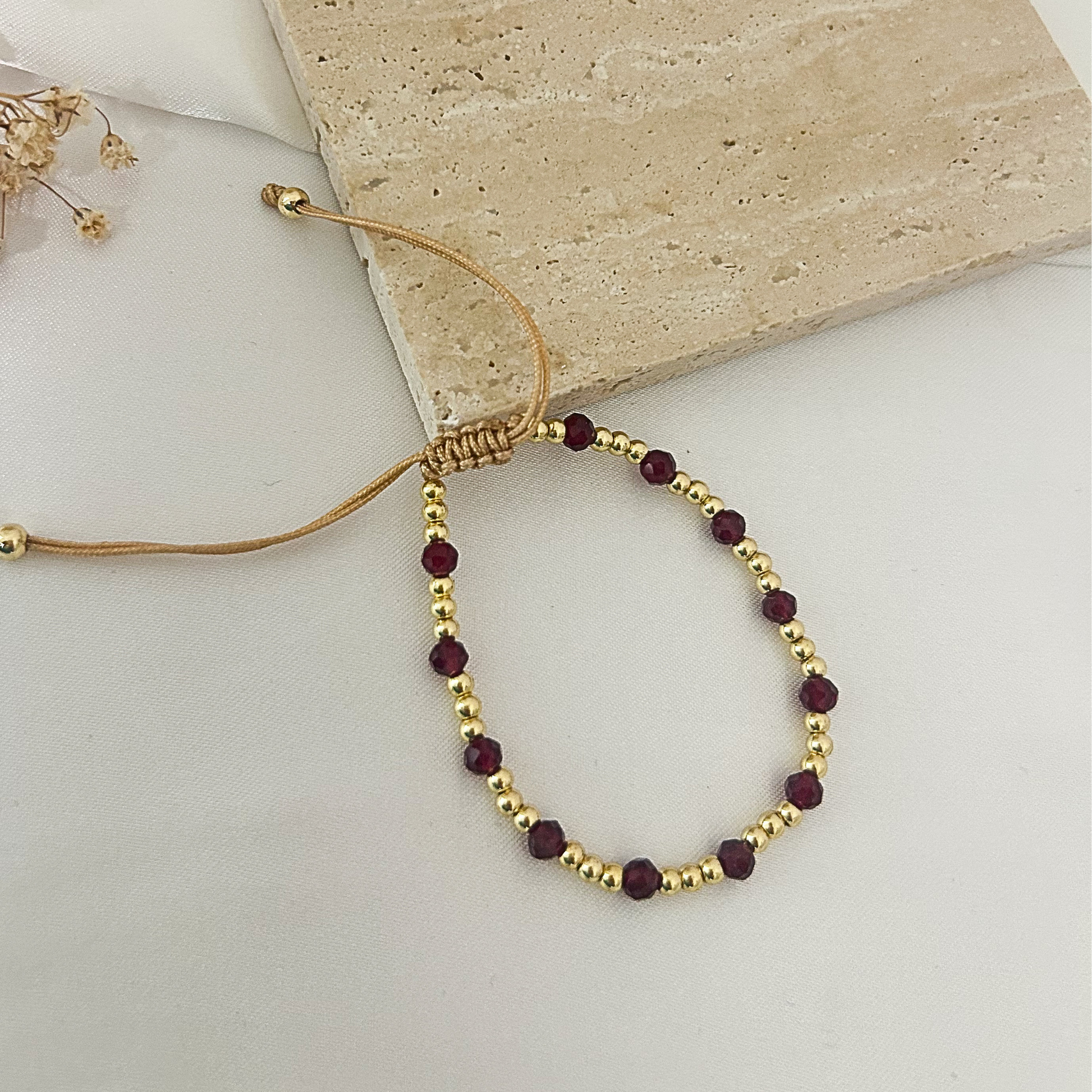 A beaded adjustable cord bracelet with gold-toned spacer beads and dark red faceted beads, laid on a light surface beside a beige stone tile.