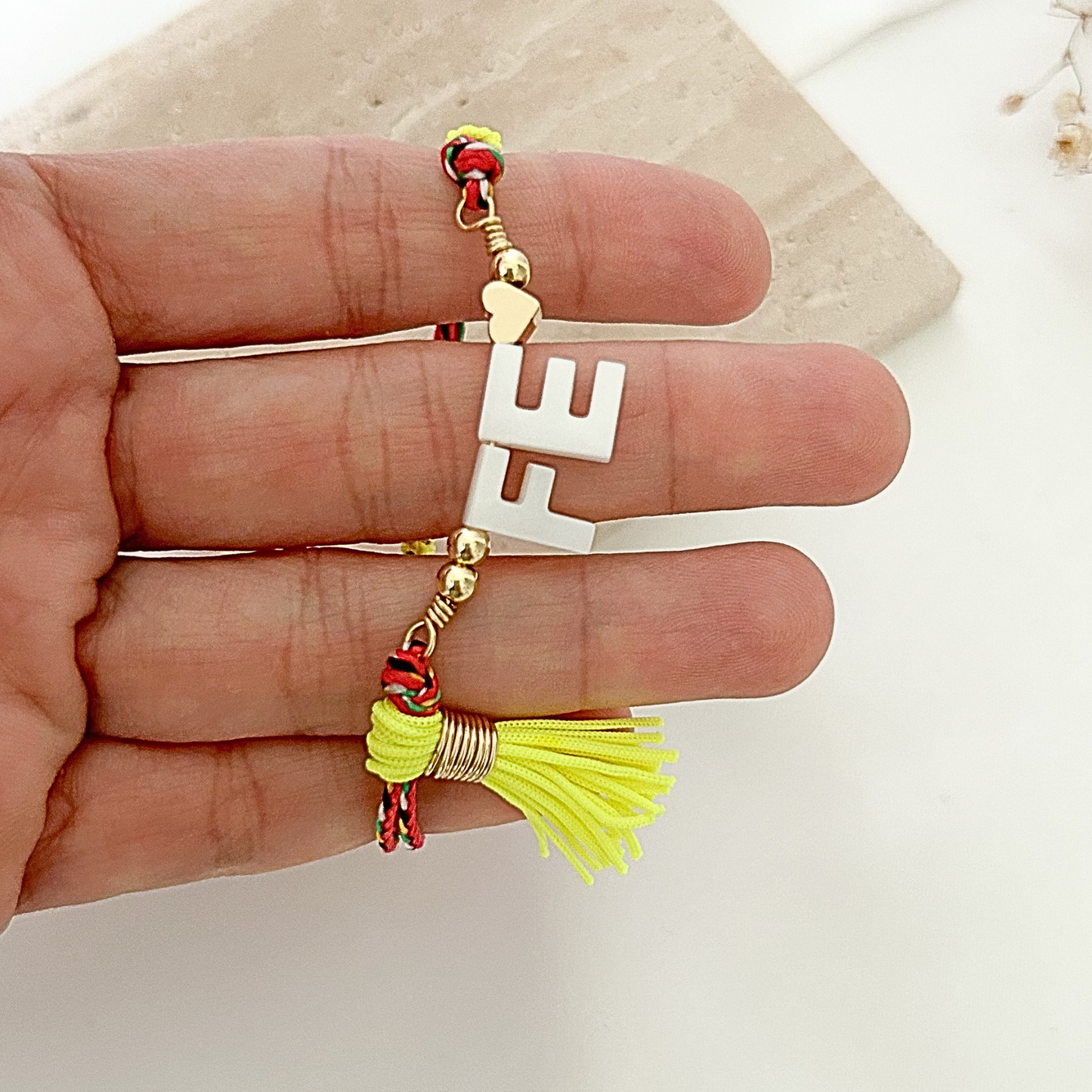 Close-up of a handmade faith bracelet with white “FE” lettering, gold-tone beads, multicolor braided cord, and a vibrant yellow tassel charm.