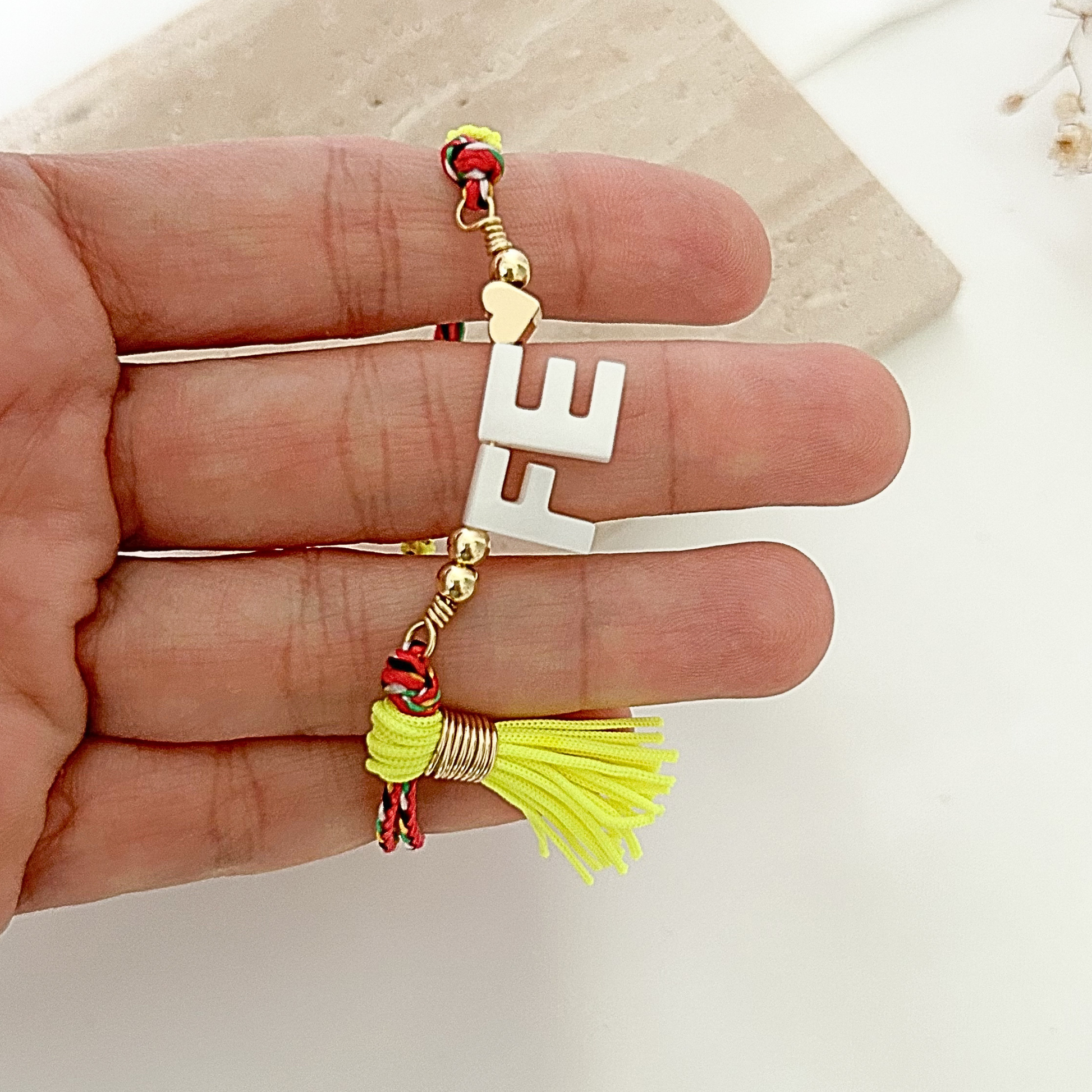 Close-up of a handmade faith bracelet with white “FE” lettering, gold-tone beads, multicolor braided cord, and a vibrant yellow tassel charm.
