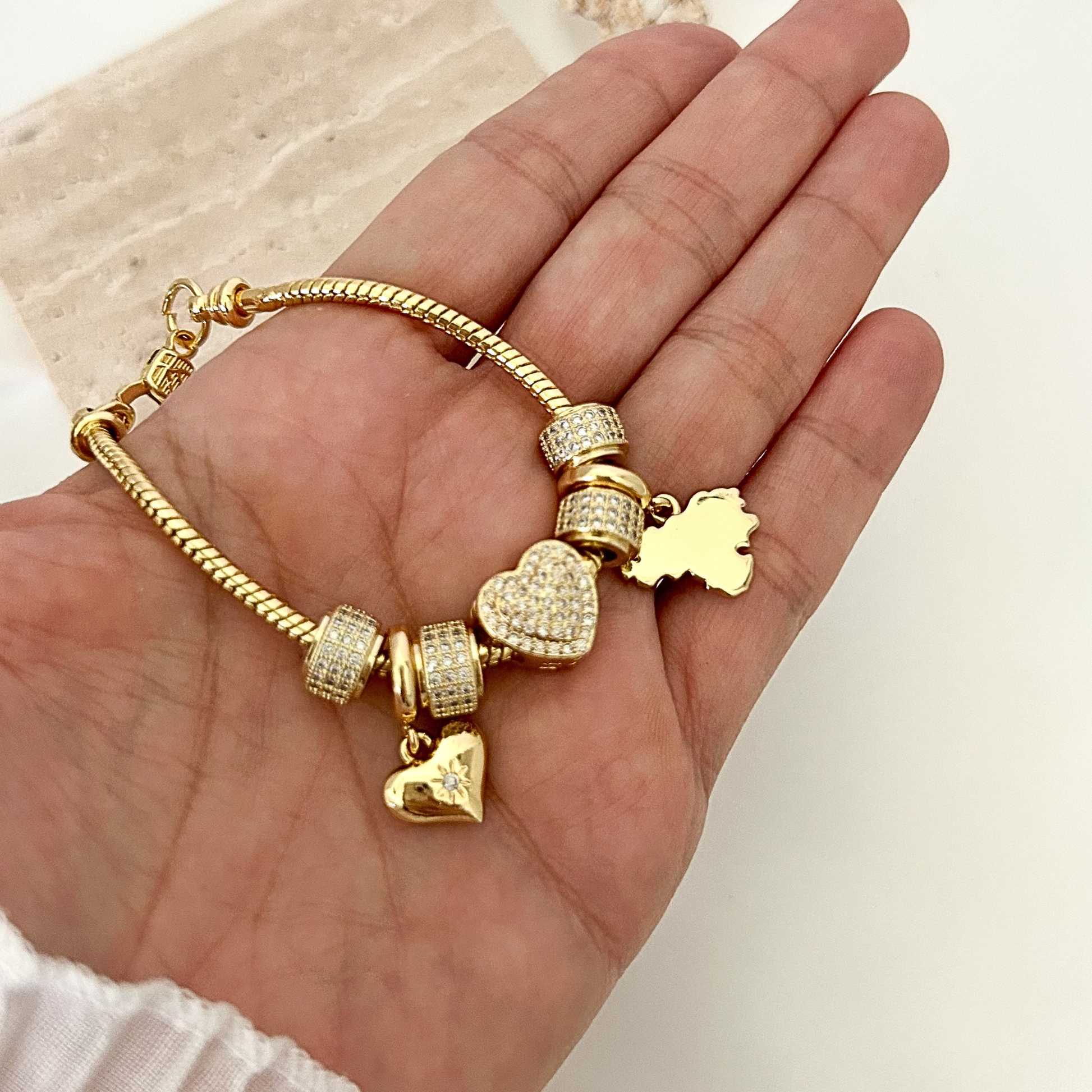 Gold-tone charm bracelet with sparkling pavé accents, featuring heart-shaped charms and a Venezuela map charm, displayed in the palm of a hand.