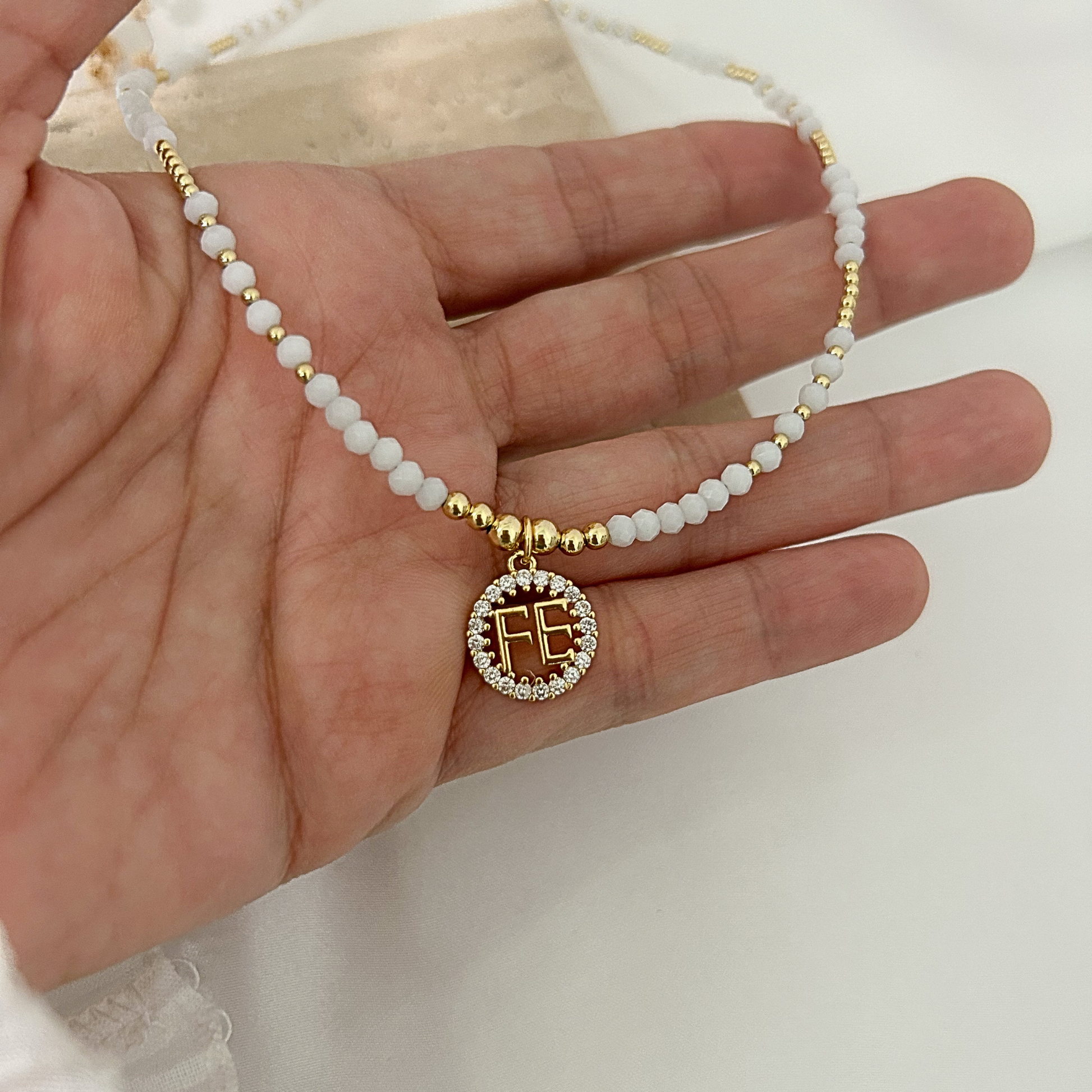 Collar de cuentas blancas y detalles dorados con medalla “FE” decorada con circonias, sostenido sobre una mano para mostrar su tamaño y acabado elegante.