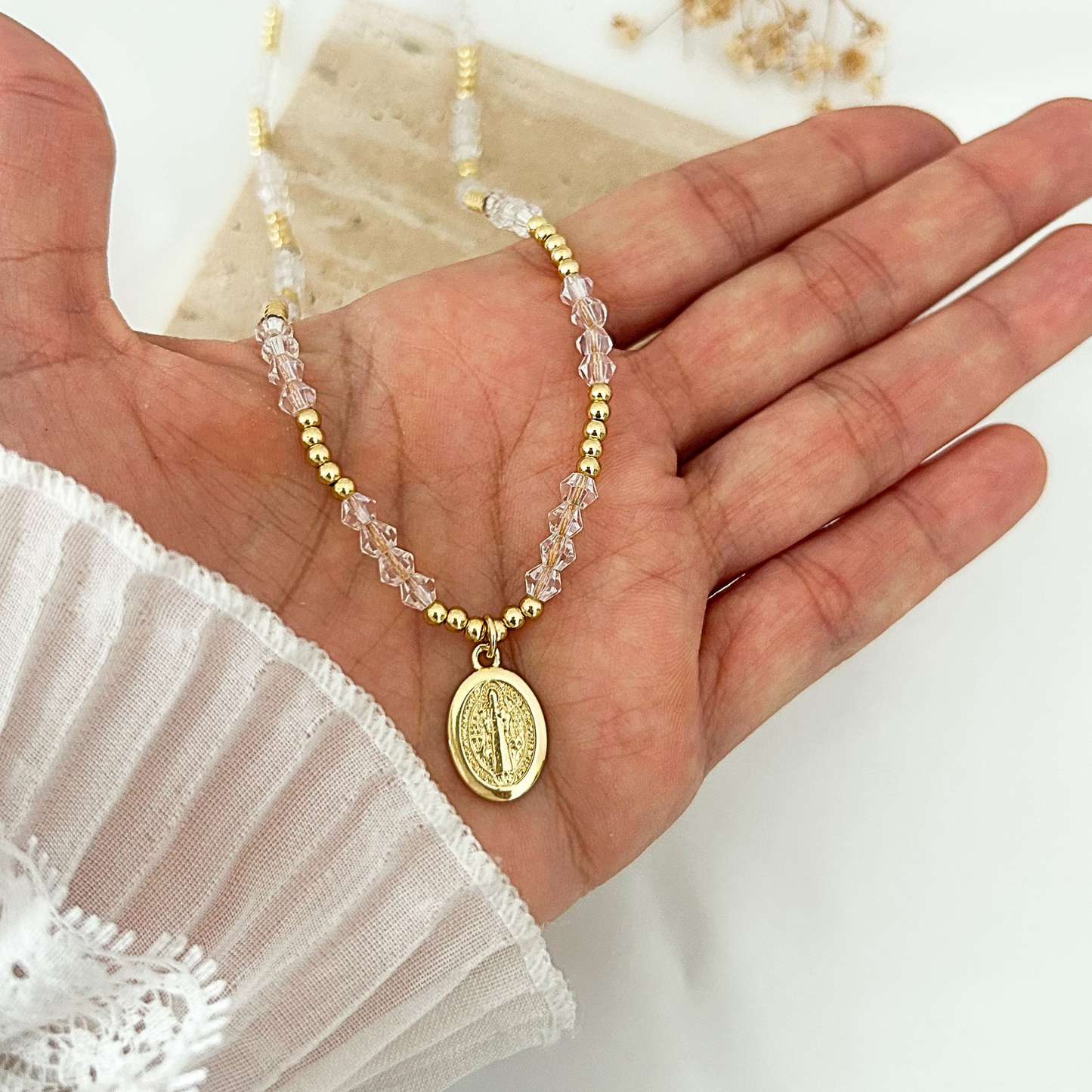 A beaded necklace with gold and clear faceted beads and a gold oval religious pendant, displayed on an open hand.