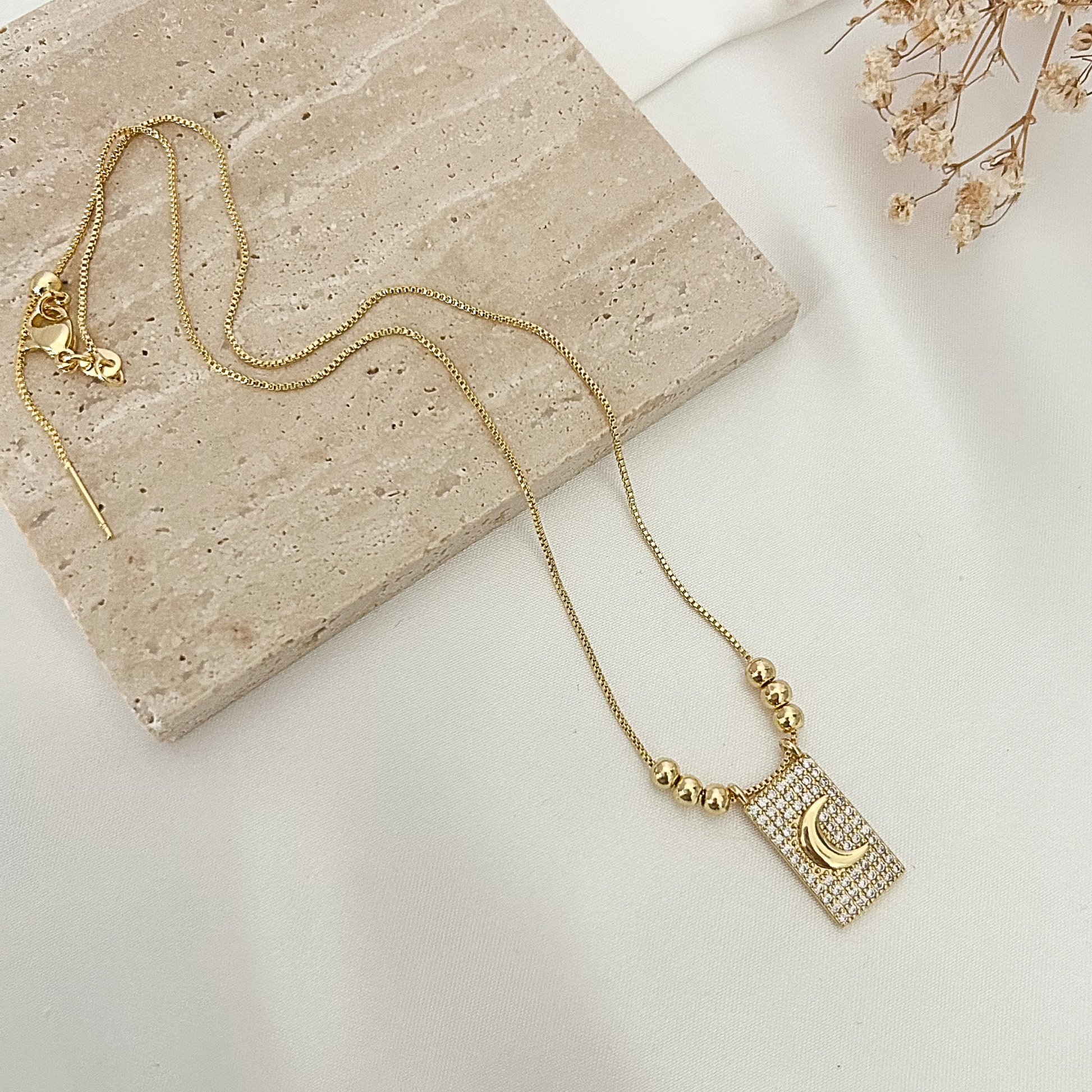Gold-tone adjustable necklace featuring a sparkling rectangular pendant with a crescent moon design, displayed on a neutral stone surface with delicate dried flowers.