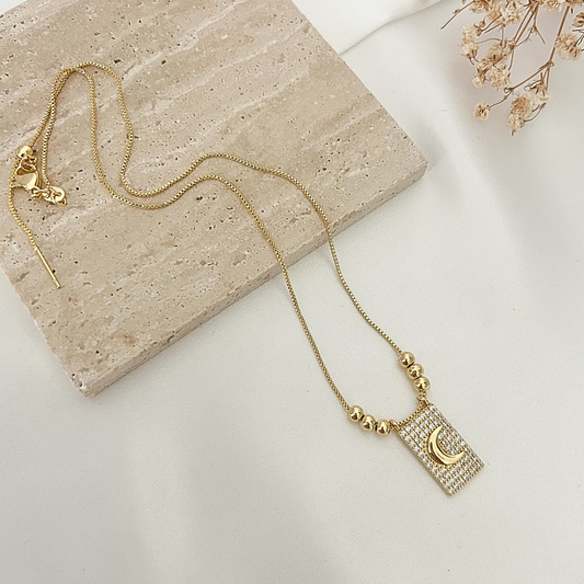 Gold-tone adjustable necklace featuring a sparkling rectangular pendant with a crescent moon design, displayed on a neutral stone surface with delicate dried flowers.