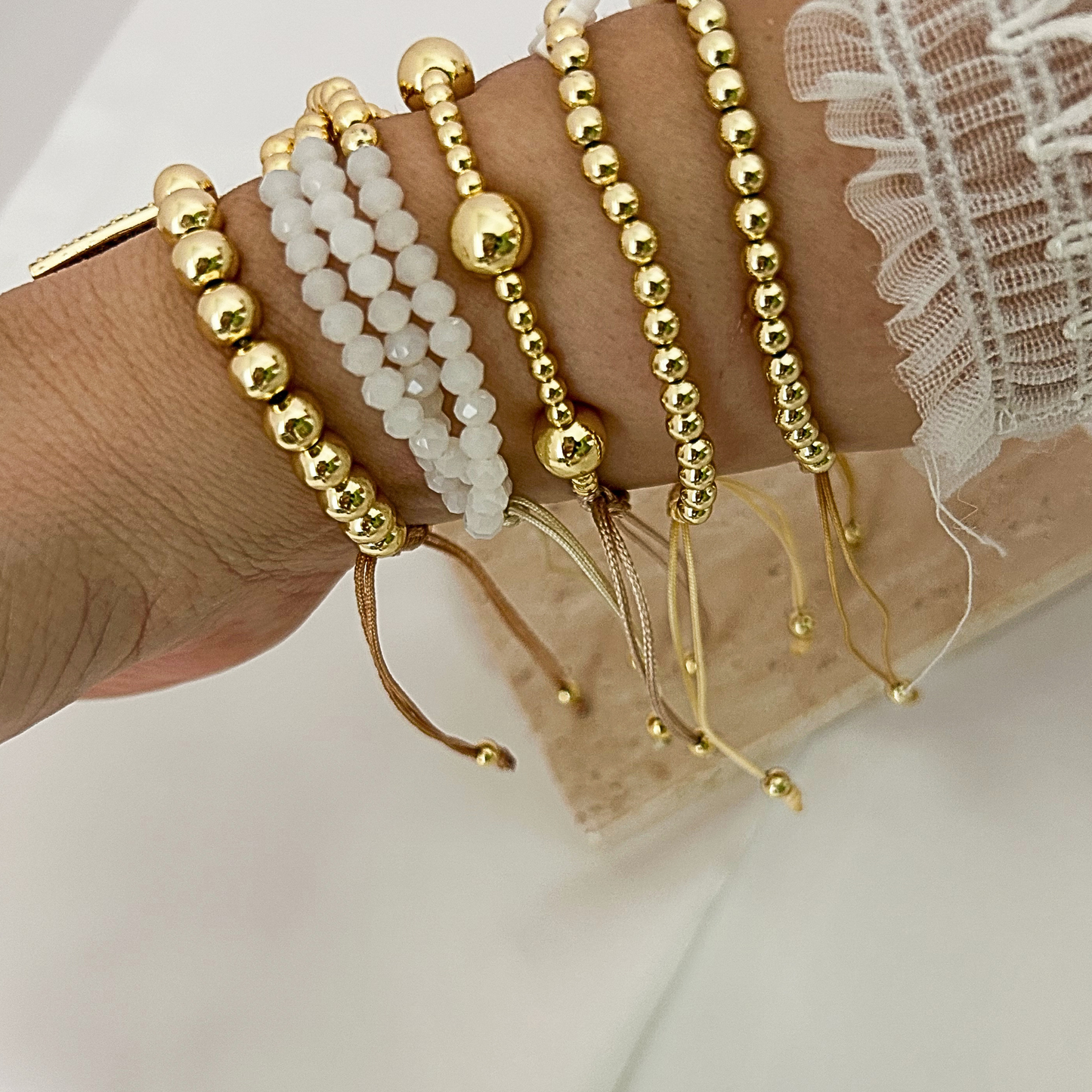 A close-up of a person's wrist wearing multiple beaded bracelets. The bracelets include gold-tone ball-beaded strands, white pearl-like beads, and thin gold cord bracelets, shown against a soft light background with a lace sleeve visible.