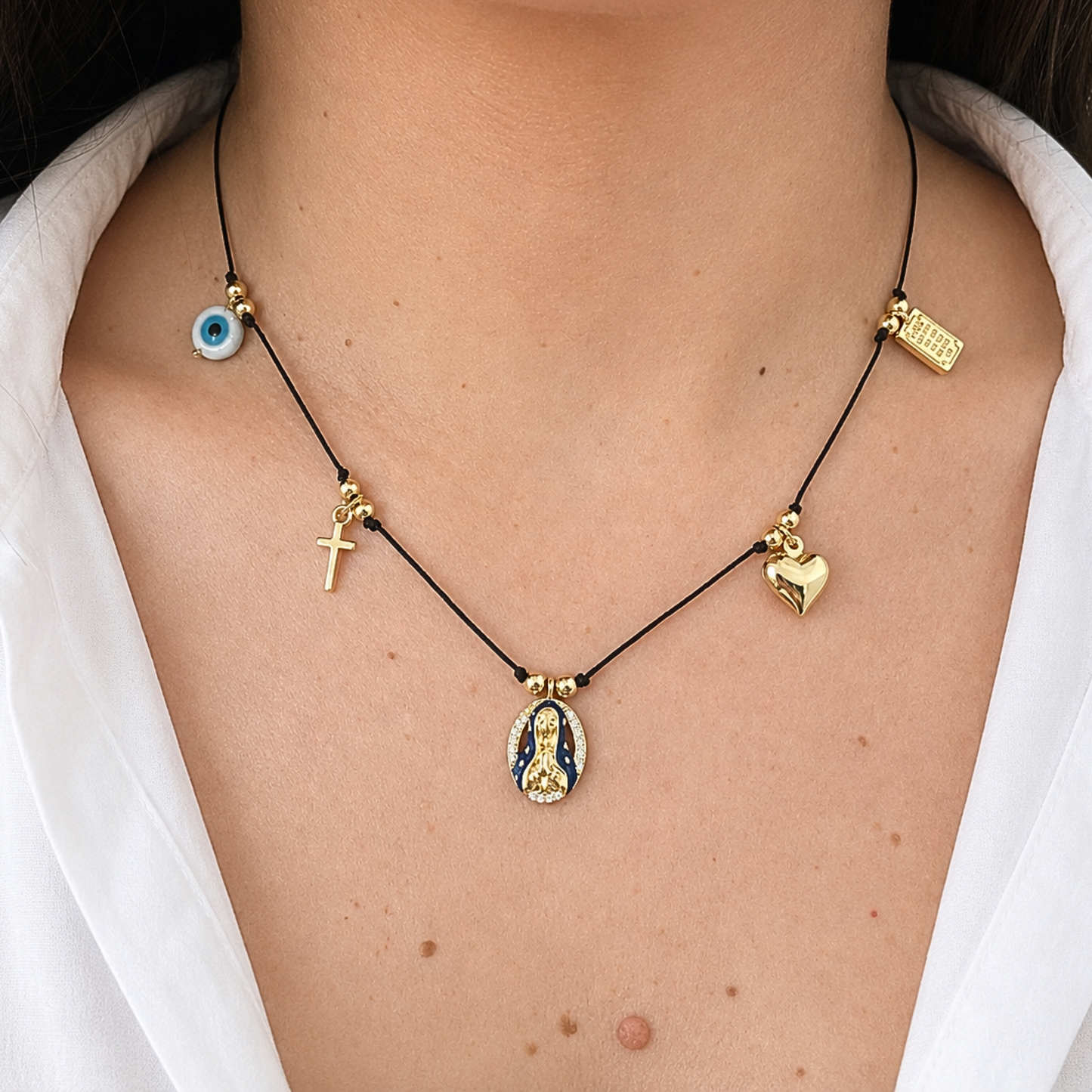 Elegant black cord necklace with meaningful religious and protective symbols, combining a gold cross, evil eye charm, heart pendant, and Virgin Mary centerpiece.