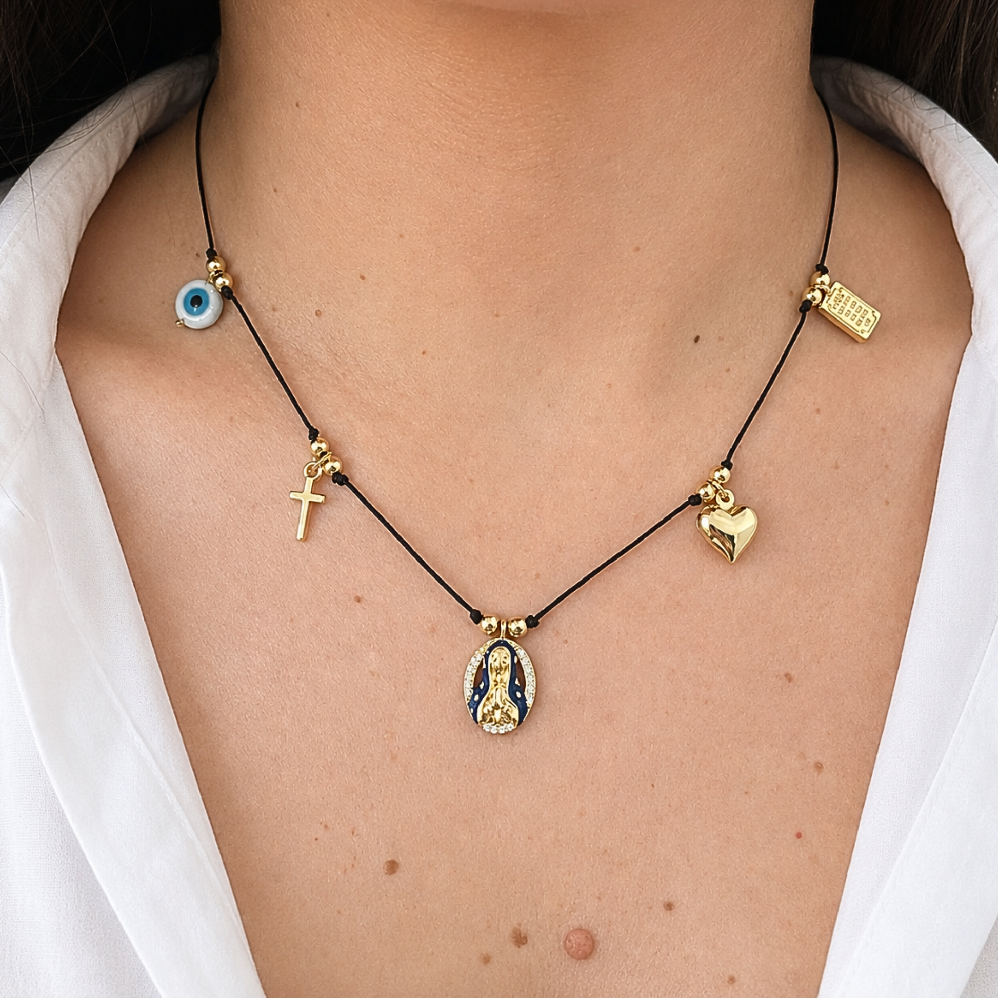 Elegant black cord necklace with meaningful religious and protective symbols, combining a gold cross, evil eye charm, heart pendant, and Virgin Mary centerpiece.