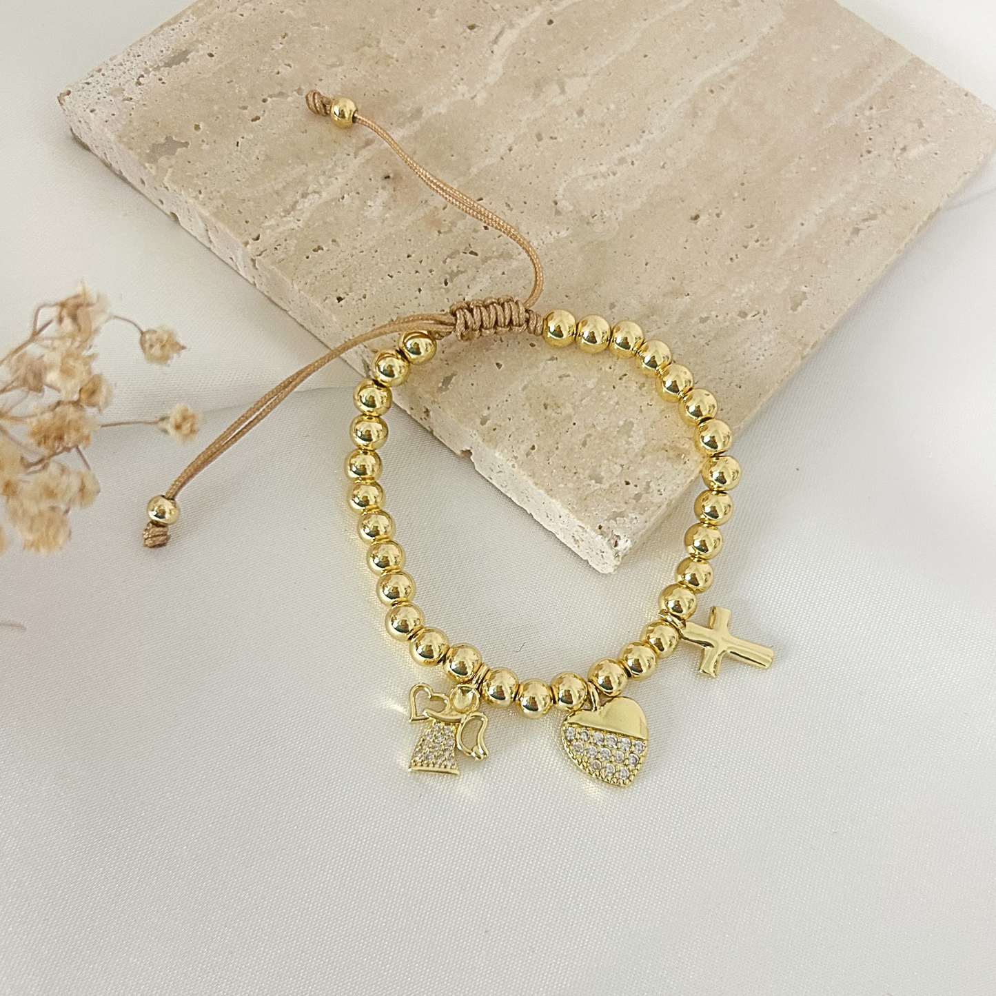A gold beaded stretch bracelet with angel and heart charms, displayed on a beige stone tile with dried flowers.