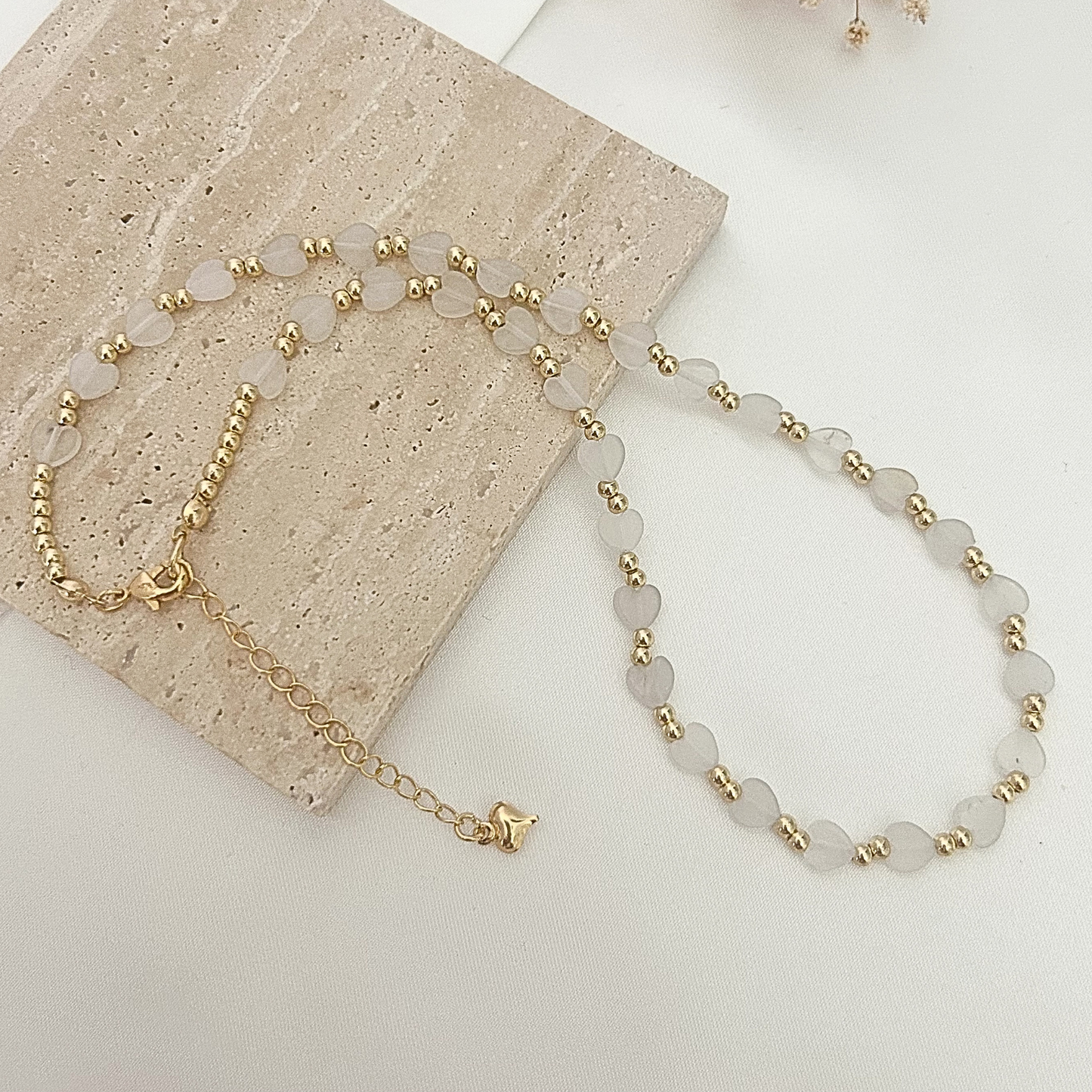 Delicate necklace featuring frosted heart-shaped beads paired with gold-tone spacer beads, displayed on a neutral stone surface.